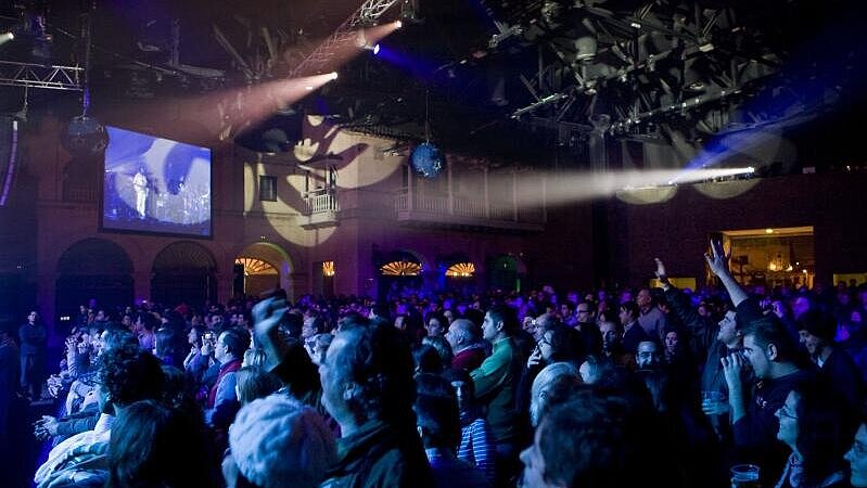 Izquierda: Concierto nocturno en interior con público bajo luces azules y focos en el escenario.